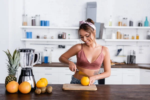 Jovem dona de casa cortando laranja perto de frutas e liquidificador na mesa da cozinha — Fotografia de Stock