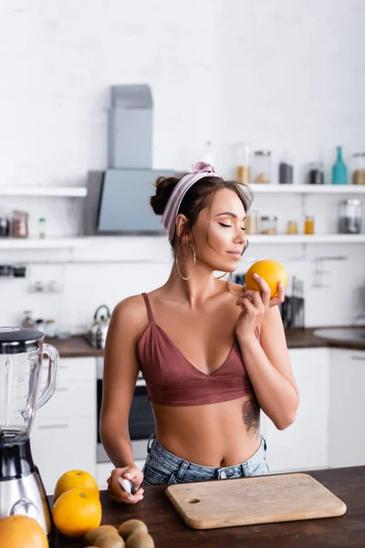 Enfoque selectivo de ama de casa oliendo naranja cerca de frutas y licuadora en la cocina - foto de stock