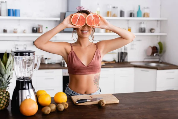 Enfoque selectivo de la mujer joven sosteniendo mitades de pomelo cerca de los ojos mientras cocina batido en la cocina - foto de stock