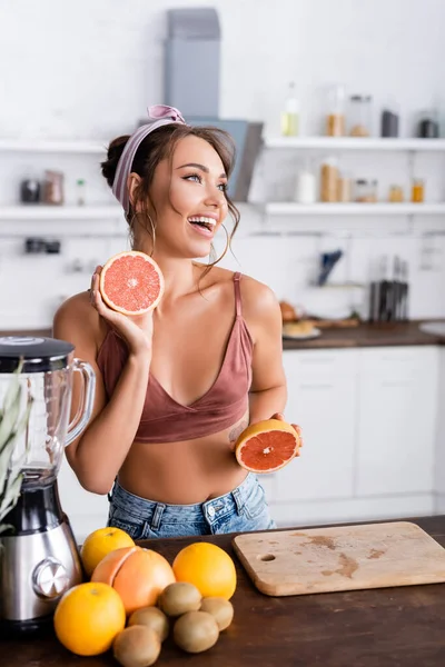 Selektiver Fokus der Hausfrau, die die Hälfte der Grapefruit in der Nähe des Mixers und der Früchte auf dem Küchentisch hält — Stockfoto