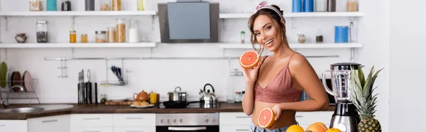 Panoramic crop of young woman looking at camera while holding halves of grapefruit near blender at home — Stock Photo