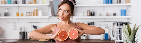 Panoramaorientierung einer brünetten Frau, die die Hälfte der Grapefruit zu Hause hält — Stockfoto
