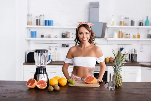 Jeune femme regardant la caméra près des fruits et mélangeur sur la table — Photo de stock