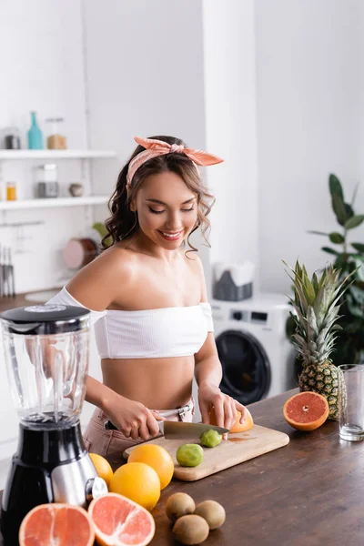 Selective focus of housewife cutting kiwi near fruits and blender in kitchen — Stock Photo