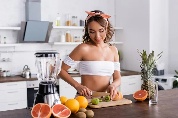 Foco seletivo de mulher jovem corte kiwi enquanto prepara smoothie na cozinha — Fotografia de Stock