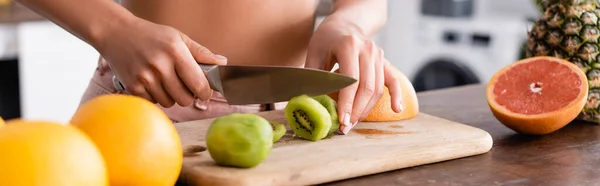 Panoramic shot of young woman cutting kiwi near fruits — Stock Photo