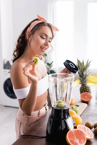 Selective focus of young woman putting kiwi in blender near fruits on table — Stock Photo