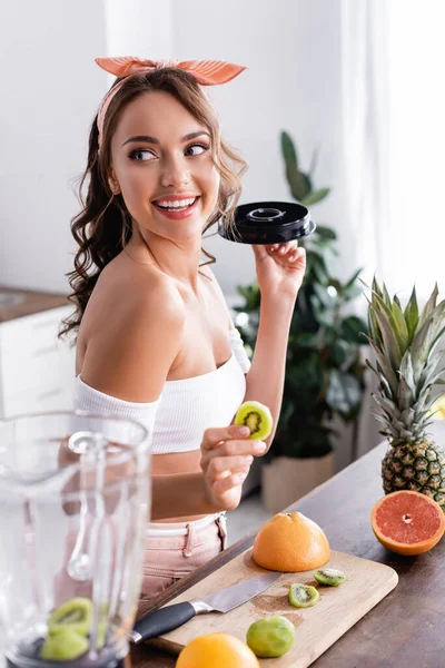 Concentration sélective de la jeune femme tenant une tranche de kiwi près des fruits et mélangeur — Photo de stock