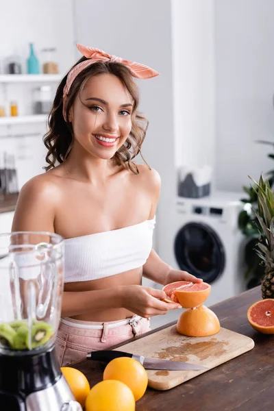 Селективное внимание домохозяйки, держащей половину грейпфрута рядом с фруктами и блендером дома — стоковое фото