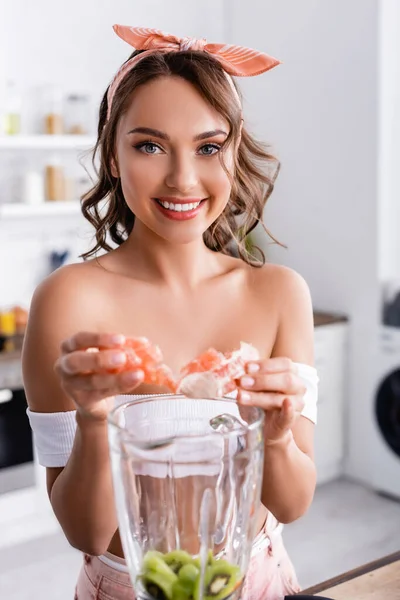 Selektiver Fokus der Hausfrau, die in die Kamera schaut, während sie Früchte im Mixer hinzufügt — Stockfoto