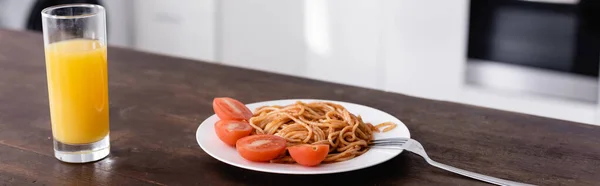 Foto panorámica de sabrosos espaguetis y vaso de jugo de naranja en la mesa de la cocina - foto de stock