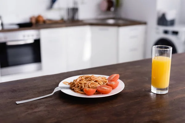 Deliciosos espaguetis y tomate en plato cerca de un vaso de jugo de naranja en la mesa de la cocina - foto de stock
