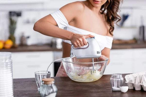 Vue recadrée de la jeune femme mélangeant les ingrédients avec un mélangeur pendant la cuisson sur la table de cuisine — Photo de stock