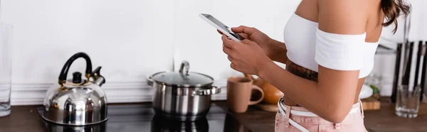 Panorâmica tiro de mulher jovem usando smartphone perto fogão na cozinha — Fotografia de Stock