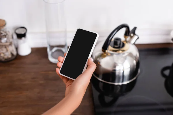 Vista recortada de la mujer sosteniendo teléfono inteligente con pantalla en blanco cerca de hervidor de agua en la estufa - foto de stock