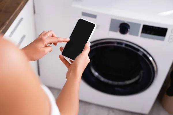 Vista cortada de mulher jovem usando smartphone perto da máquina de lavar roupa automatizada — Fotografia de Stock