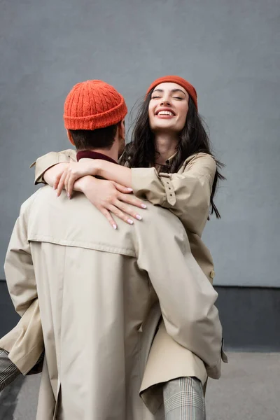 Vue arrière de l'homme tenant dans les bras femme joyeuse en bonnet chapeau et trench coat — Photo de stock