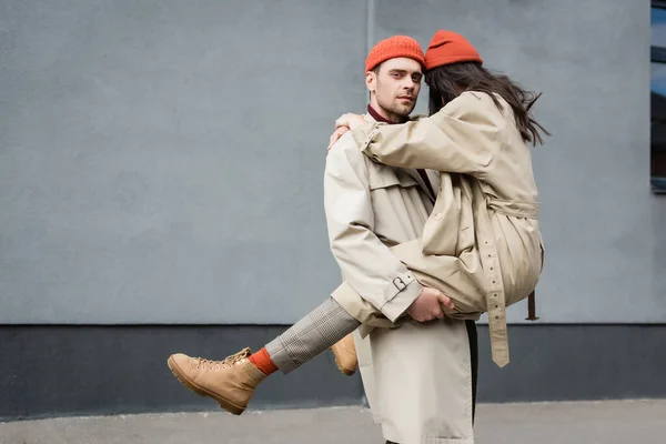 Stylish man holding in arms woman in beanie hat and trench coat — Stock Photo