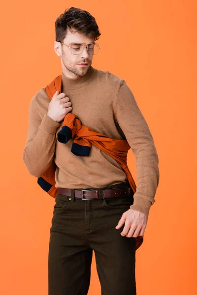Uomo elegante in occhiali che toccano maglione e guardando lontano isolato su arancione — Foto stock