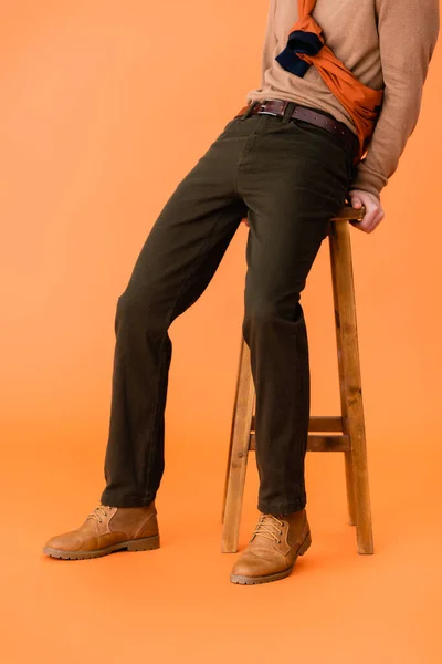 Cropped view of trendy man in autumn outfit sitting on wooden stool on orange — Stock Photo