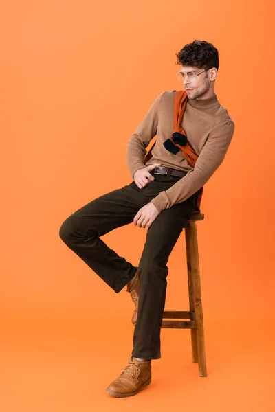 Stylish man in autumn outfit and glasses sitting on wooden stool on orange — Stock Photo