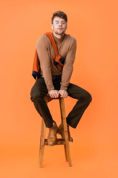 Homme à la mode en tenue d'automne et lunettes assises sur tabouret en bois sur orange — Photo de stock