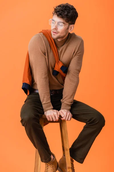 Homem elegante em roupa de outono e óculos sentados em banquinho de madeira e olhando para longe isolado em laranja — Fotografia de Stock