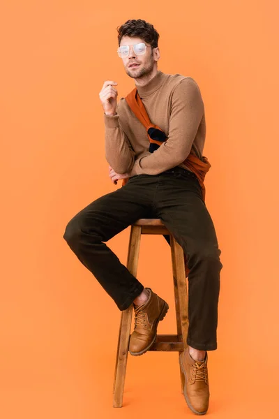 Hombre pensativo en traje de otoño y gafas sentados en taburete de madera en naranja - foto de stock