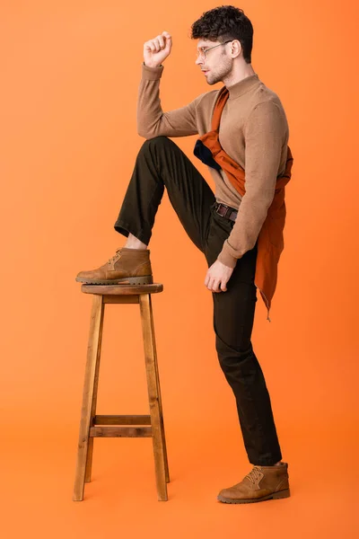 Side view of stylish man in autumn outfit and glasses standing with leg on wooden stool on orange — Stock Photo