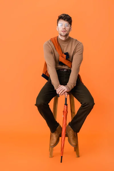 Fashionable man in autumn outfit and glasses holding umbrella and sitting on wooden stool on orange — Stock Photo