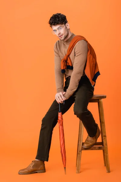 Hombre en traje de otoño y gafas sosteniendo paraguas mientras está sentado en taburete de madera en naranja - foto de stock