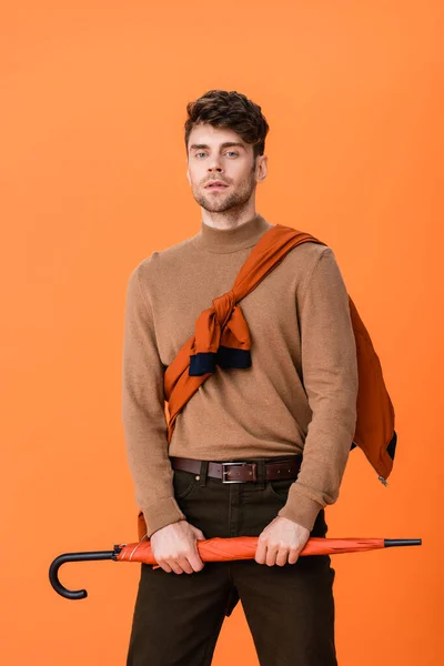 Hombre con estilo en traje de otoño que sostiene el paraguas mientras mira a la cámara aislada en naranja - foto de stock