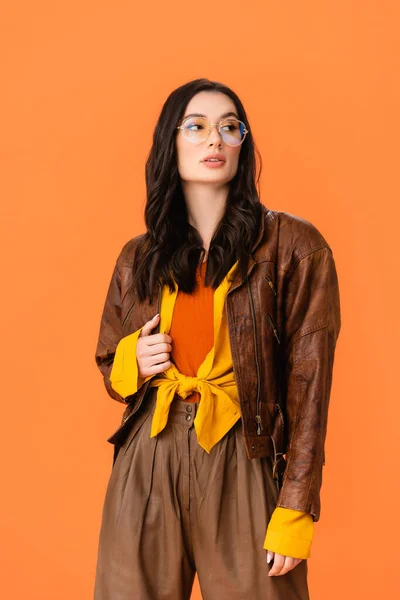 Mujer con estilo en traje de otoño y gafas tocando chaqueta y mirando hacia otro lado aislado en naranja - foto de stock