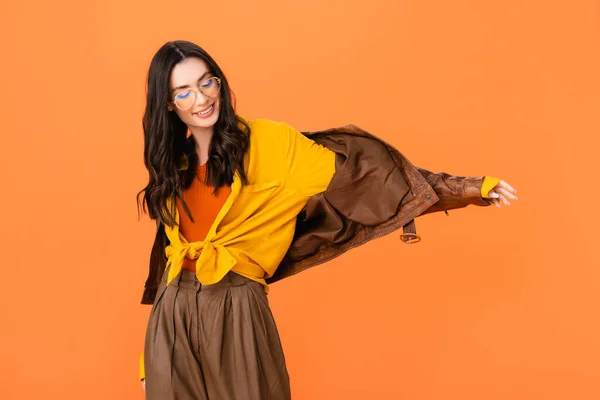 Elegante donna in occhiali e giacca di pelle in piedi con mano tesa isolata su arancione — Foto stock