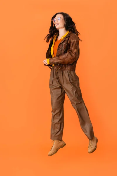 Longitud completa de la mujer joven en traje de otoño y gafas levitando en naranja - foto de stock