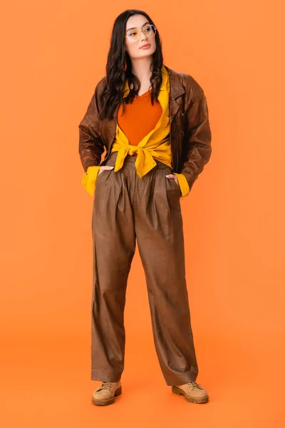 Longitud completa de la mujer joven en traje de otoño de pie con las manos en los bolsillos en naranja - foto de stock