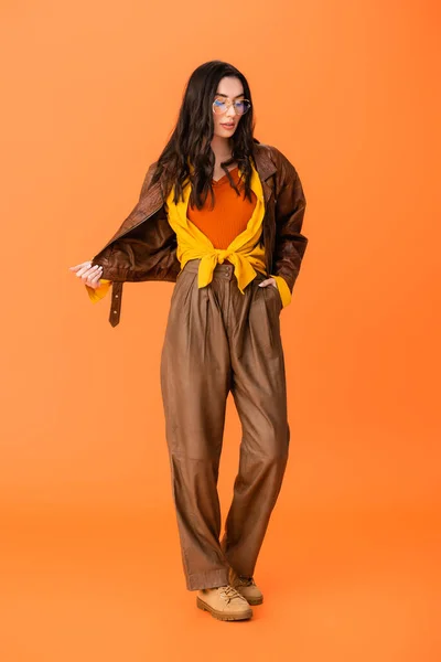 Full length of young woman in autumn outfit standing with hand in pocket and touching jacket on orange — Stock Photo