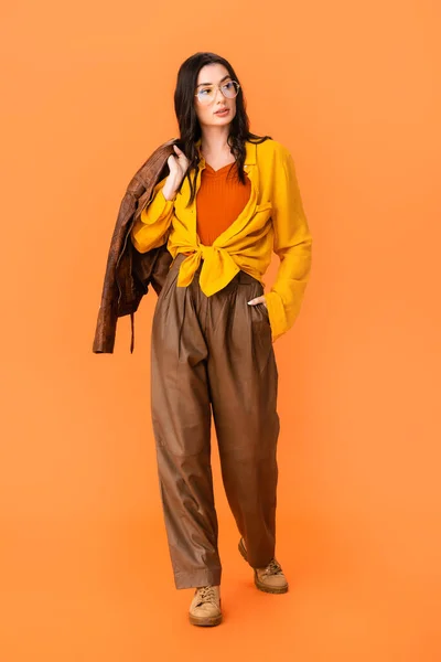 Comprimento total da jovem mulher em roupa de outono segurando jaqueta de couro e de pé com a mão no bolso em laranja — Fotografia de Stock