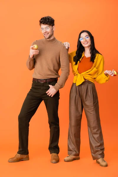 Piena lunghezza di coppia alla moda in bicchieri e abbigliamento autunnale che tiene mele su arancione — Foto stock