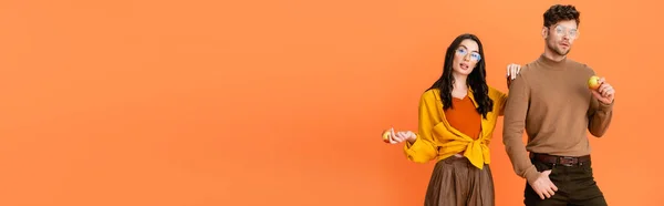 Panoramic crop of trendy couple in glasses and autumn outfit holding apples isolated on orange — Stock Photo