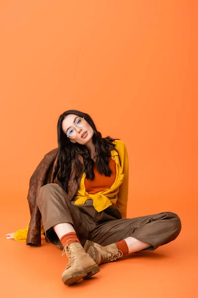 Donna di tendenza in occhiali e abbigliamento autunnale e giacca di pelle seduta su arancione — Foto stock