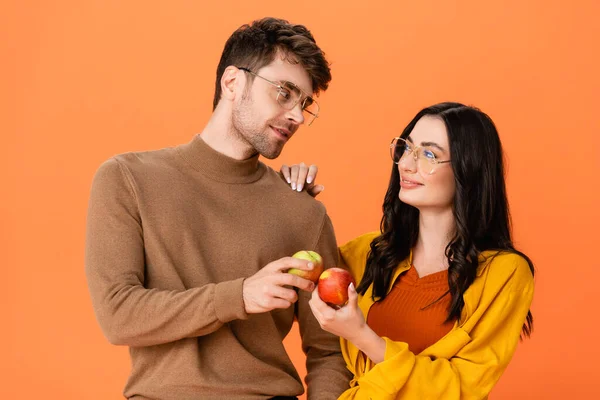 Pareja de moda en gafas y traje de otoño sosteniendo manzanas mientras se miran aislados en naranja - foto de stock