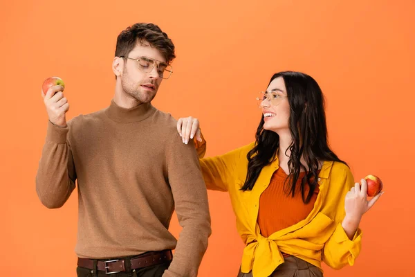 Elegante pareja en gafas y traje de otoño sosteniendo manzanas mientras se miran aislados en naranja - foto de stock