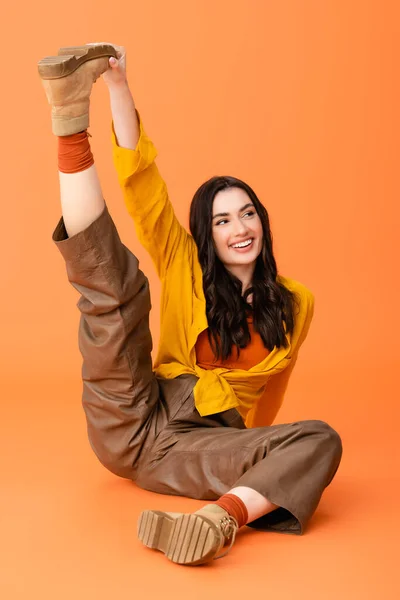 Mujer de moda en traje de otoño estiramiento de la pierna y sentado en naranja - foto de stock