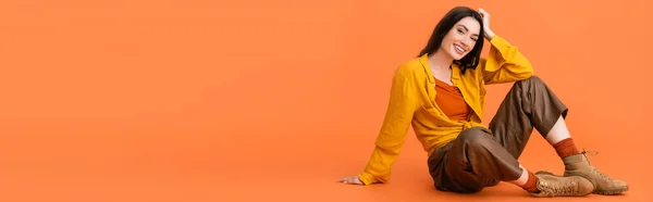 Concepto panorámico de morena y mujer de moda en traje de otoño sentado en naranja - foto de stock