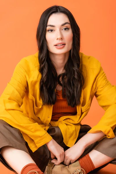 Brunette young woman in autumn outfit looking at camera and sitting isolated on orange — Stock Photo