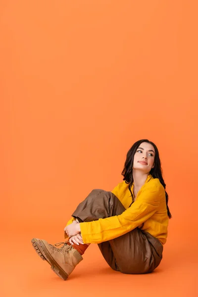 Jovem morena em roupa de outono e botas olhando embora enquanto sentado isolado em laranja — Fotografia de Stock