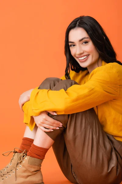 Joyful young woman in autumn outfit and boots looking at camera while sitting isolated on orange — Stock Photo