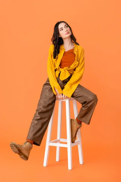 Piena lunghezza di giovane donna in abito autunnale e stivali seduta su sgabello bianco su arancione — Foto stock