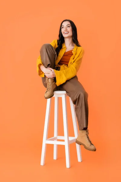 Pleased woman in autumn outfit sitting on white stool on orange — Stock Photo